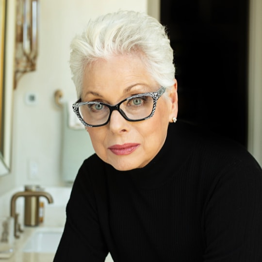mature white woman with white/gray hair wearing glasses and a black long sleeve shirt looking directly at the camera